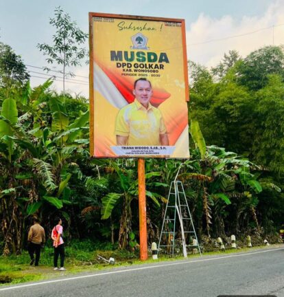 Large yellow campaign billboard on a wooden post near a road, featuring a man in a yellow shirt and bold black text for MUSDA DPD GOLKAR, surrounded by green plants and a ladder nearby.