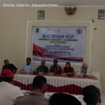 Desa meeting panel seated behind a long table with a large banner in the background reading musyawarah desa and desa kokosan 7 mei 2026 indonesian village council event