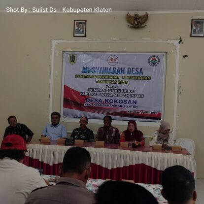 Desa meeting panel seated behind a long table with a large banner in the background reading musyawarah desa and desa kokosan 7 mei 2026 indonesian village council event