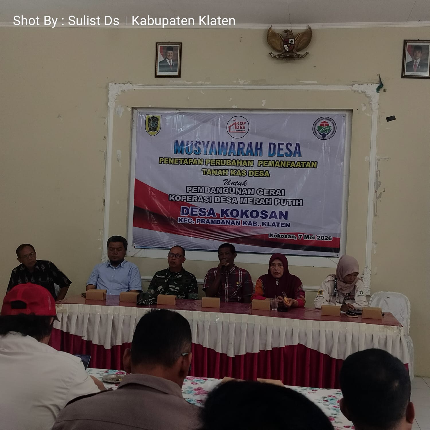 Desa meeting panel seated behind a long table with a large banner in the background reading musyawarah desa and desa kokosan 7 mei 2026 indonesian village council event