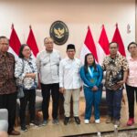 Group of eight people standing in a room with indonesian flags and a government seal behind them posing for a formal photo