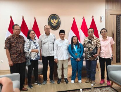 Group of eight people standing in a room with Indonesian flags and a government seal behind them, posing for a formal photo.