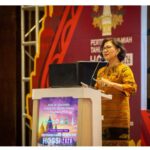 Female speaker in an orange batik dress delivering a lecture at a conference podium with a laptop and microphone close by