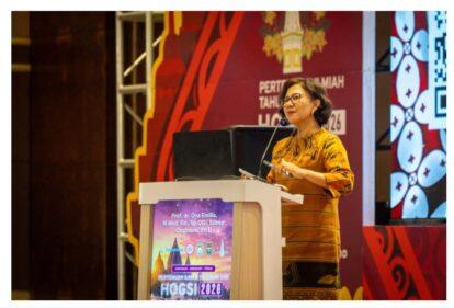 Female speaker in an orange batik dress delivering a lecture at a conference podium with a laptop and microphone close by.