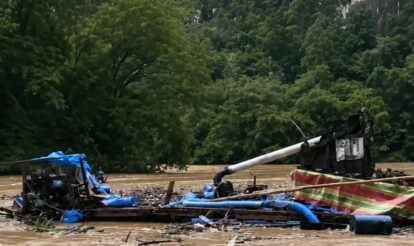 Wrecked boat and scattered debris floating in a muddy floodplain with trees in the background indicating recent flood damage