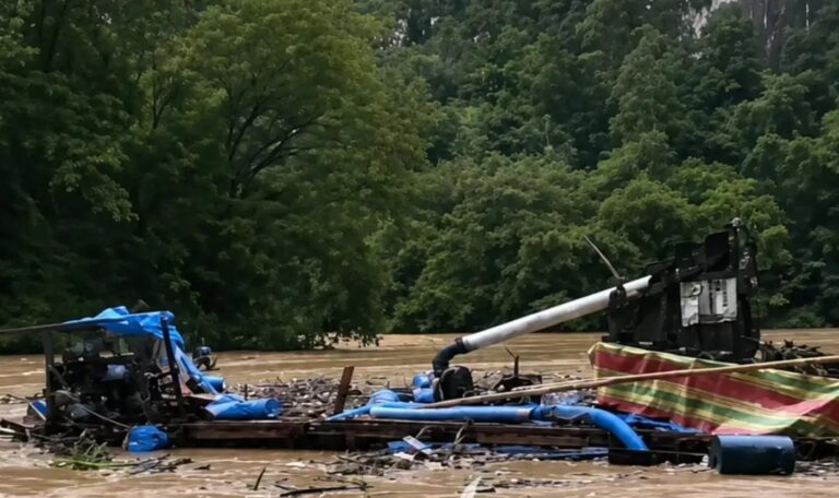 Wrecked boat and scattered debris floating in a muddy floodplain with trees in the background, indicating recent flood damage.