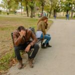Man and woman sit back to back on a park bench along a gravel path both holding their heads as if stressed with autumn trees and fallen leaves around them