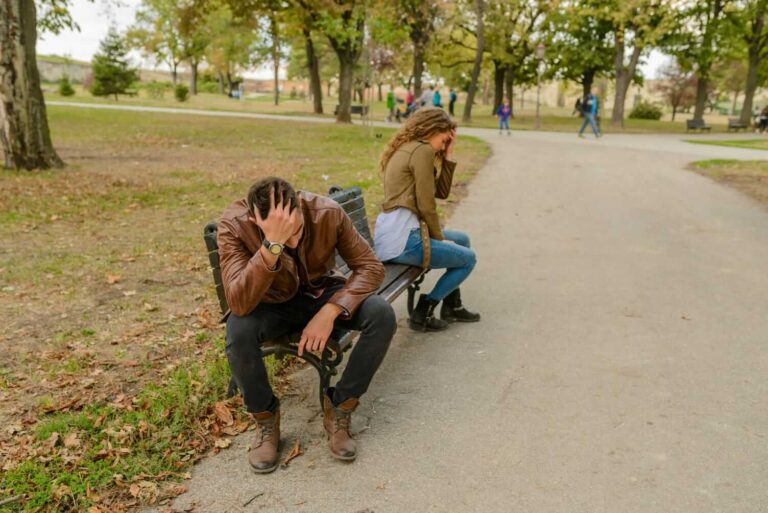 Man and woman sit back-to-back on a park bench along a gravel path, both holding their heads as if stressed, with autumn trees and fallen leaves around them.