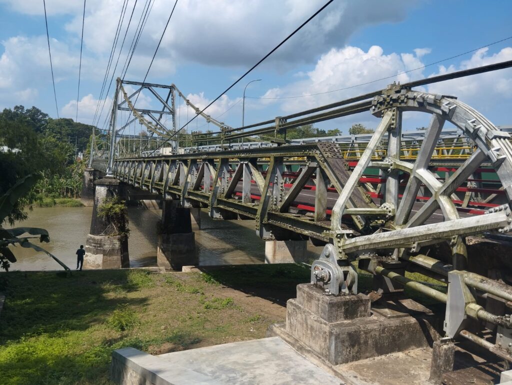 Metal truss bridge spanning a calm river with exposed stone piers under a blue sky with clouds a person stands on the riverbank nearby