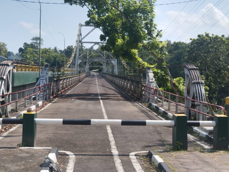 Empty metal truss bridge with red railings and barrier gate, stretching into a tree-lined background under a blue sky.