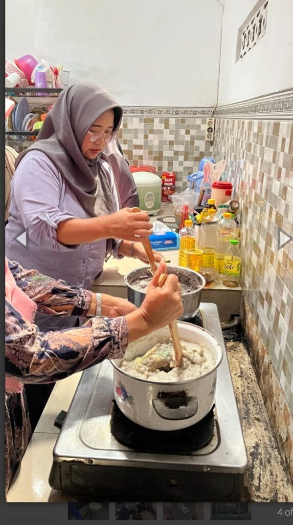Two women cooking in a small kitchen one wearing a hijab stirs a large pot while the other helps with a wooden spoon