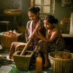 Two young girls sit in a rustic kitchen grinding spices with a large mortar and pestle