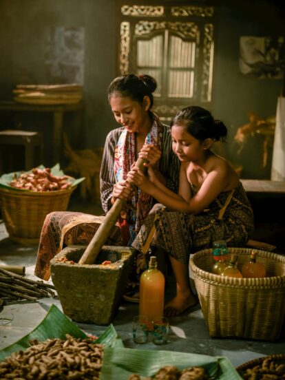 Two young girls sit in a rustic kitchen, grinding spices with a large mortar and pestle.