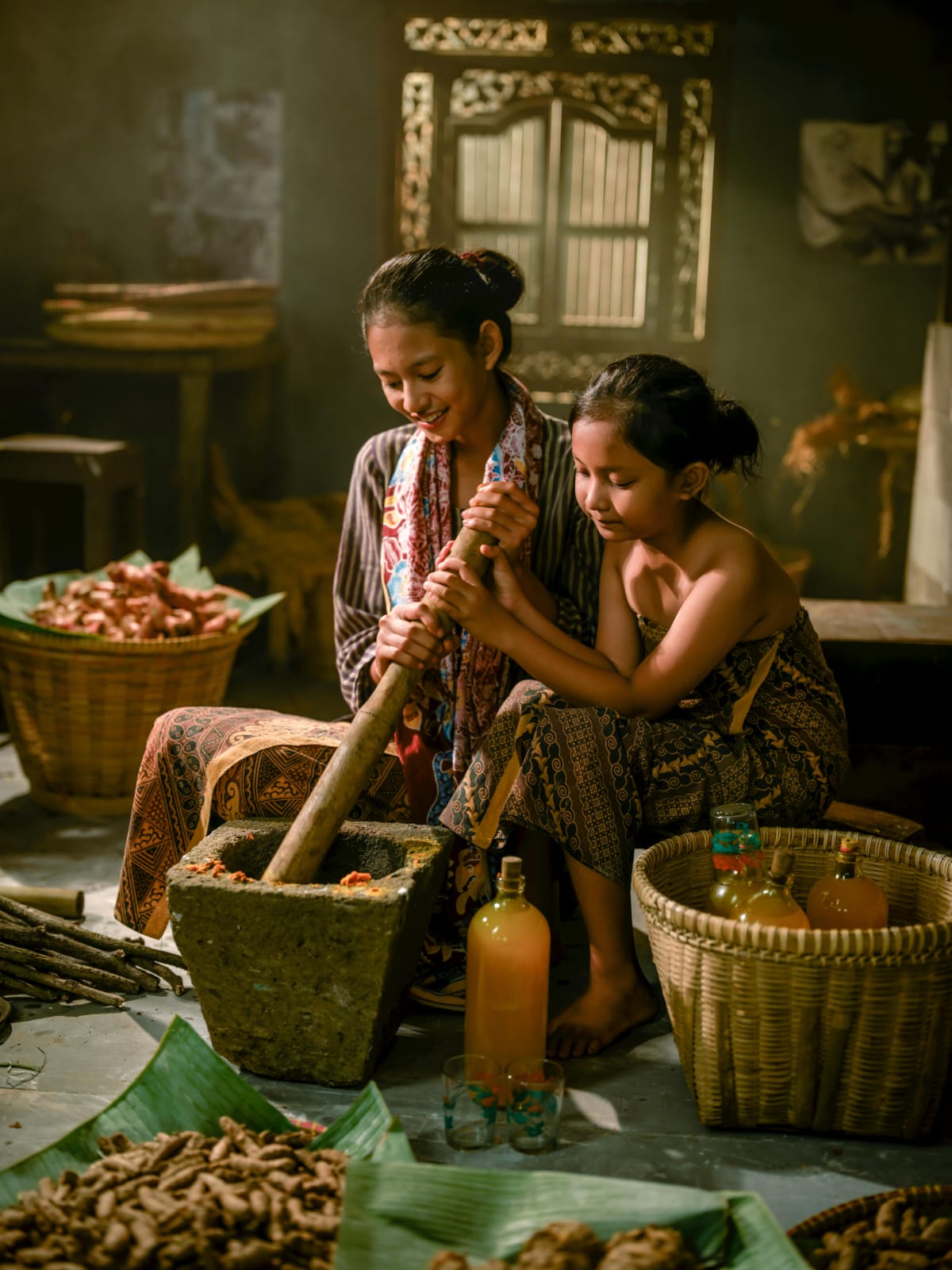 Two young girls sit in a rustic kitchen grinding spices with a large mortar and pestle