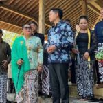 Group of people in batik and traditional attire on an outdoor stage under a thatched roof a woman in a turquoise shawl speaks into a microphone while others watch