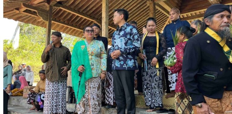 Group of people in batik and traditional attire on an outdoor stage under a thatched roof; a woman in a turquoise shawl speaks into a microphone while others watch.