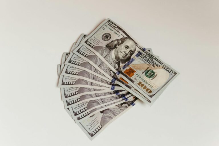 Stack of US $100 bills fanned out on a light surface, showing multiple notes spread in a semicircle.