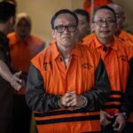 Man in an orange detainee vest with hands clasped flanked by others in similar uniforms indoors looking at the camera