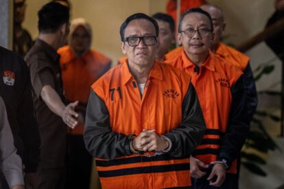 Man in an orange detainee vest with hands clasped, flanked by others in similar uniforms indoors, looking at the camera.