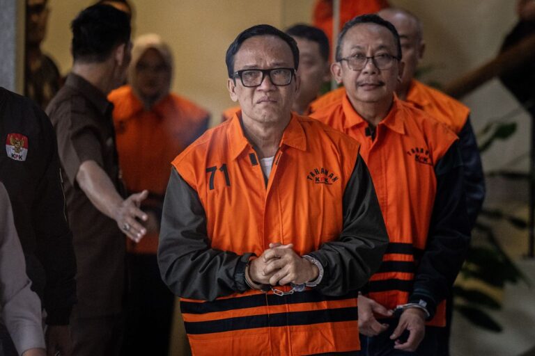 Man in an orange detainee vest with hands clasped, flanked by others in similar uniforms indoors, looking at the camera.