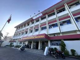 Front view of a white multi-story Indonesian government building with red accents, a flagpole displaying the Indonesian flag, and motorcycles parked outside.