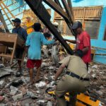 People search through rubble amid a collapsed building with wooden beams and a blue wall in the background