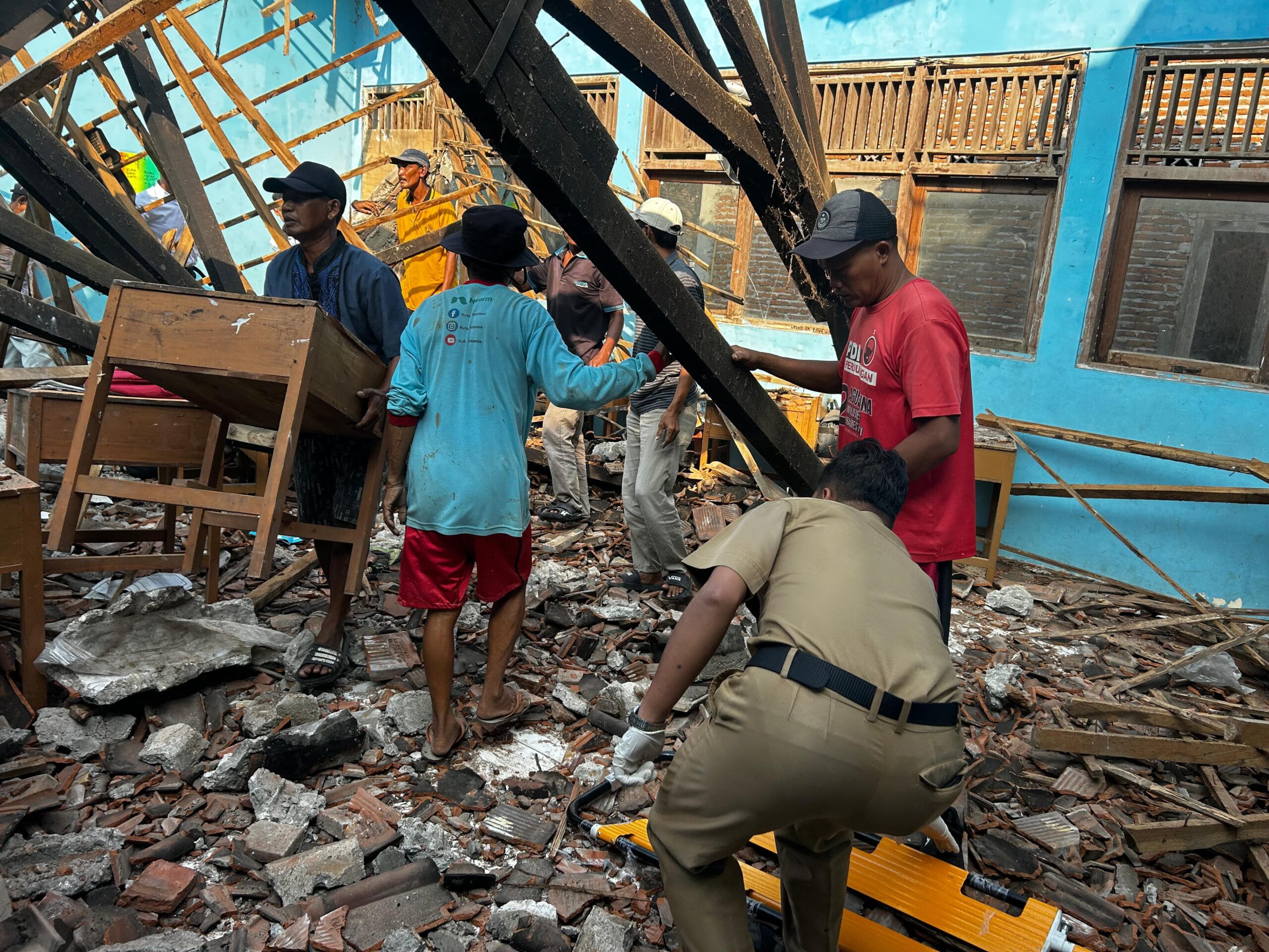 People search through rubble amid a collapsed building with wooden beams and a blue wall in the background