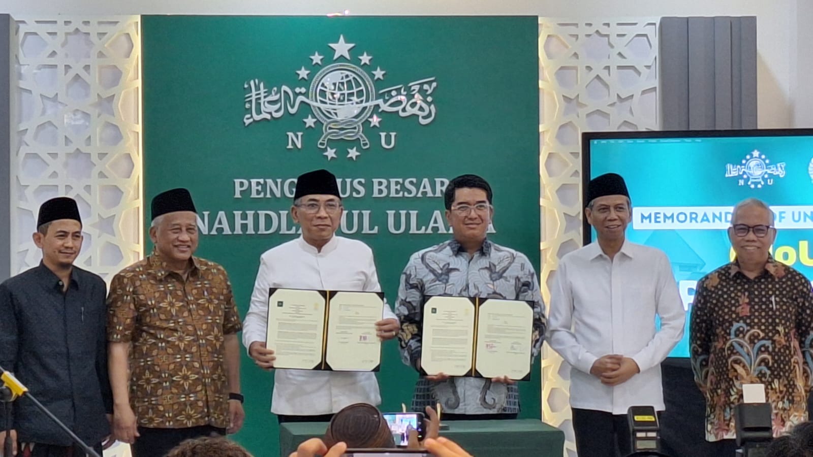 Six men on a stage hold open signed documents as a ceremony takes place with a green backdrop and attendees watching in front