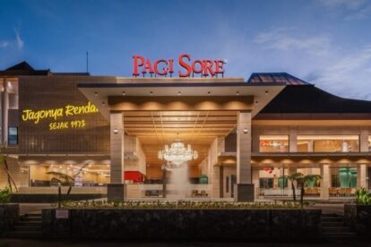 Front view of a mall entrance at dusk, with a grand chandelier fountain visible inside and bright red 'Pagi Sore' sign above.