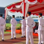 Uniformed men in white dress uniforms stand with hands behind their backs under a red and white ceremonial canopy at a stadium ceremony