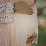 Pregnant belly cradled by hands in a pink lace dress with rings visible on fingers