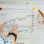 Two people review a printed stock chart pinned to a whiteboard with fingers pointing at data points and trends