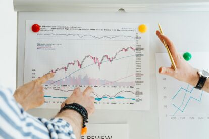 Two people review a printed stock chart pinned to a whiteboard, with fingers pointing at data points and trends.