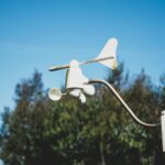 White weather vaneanemometer apparatus mounted on a pole against a clear blue sky with trees in the background