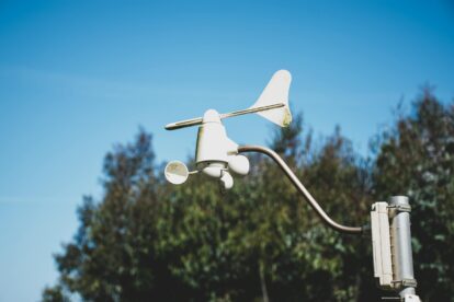 White weather vane/anemometer apparatus mounted on a pole against a clear blue sky, with trees in the background.