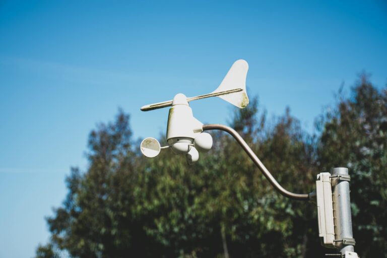 White weather vane/anemometer apparatus mounted on a pole against a clear blue sky, with trees in the background.