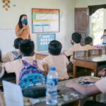 Teacher in an orange shirt and blue mask instructs a classroom students in uniform facing front the lesson poster on the wall