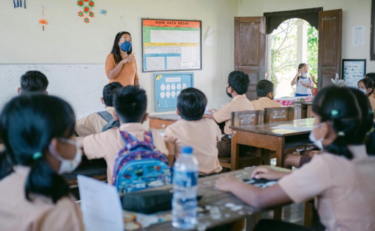 Teacher in an orange shirt and blue mask instructs a classroom, students in uniform facing front the lesson poster on the wall.