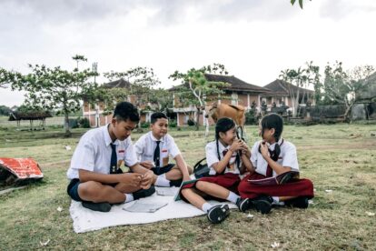 Group of Indonesian schoolchildren in white shirts and navy skirts/shorts sitting on a white blanket outdoors, studying papers with a magnifying glass, a cow in the background near brick buildings.