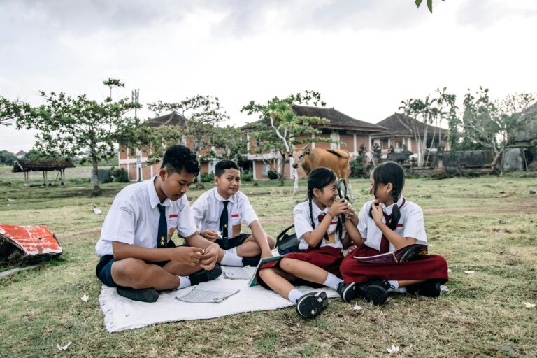 Group of Indonesian schoolchildren in white shirts and navy skirts/shorts sitting on a white blanket outdoors, studying papers with a magnifying glass, a cow in the background near brick buildings.