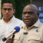 Close up of a uniformed officer speaking into a handheld microphone at a press event with a second man in a light shirt visible in the blurred background