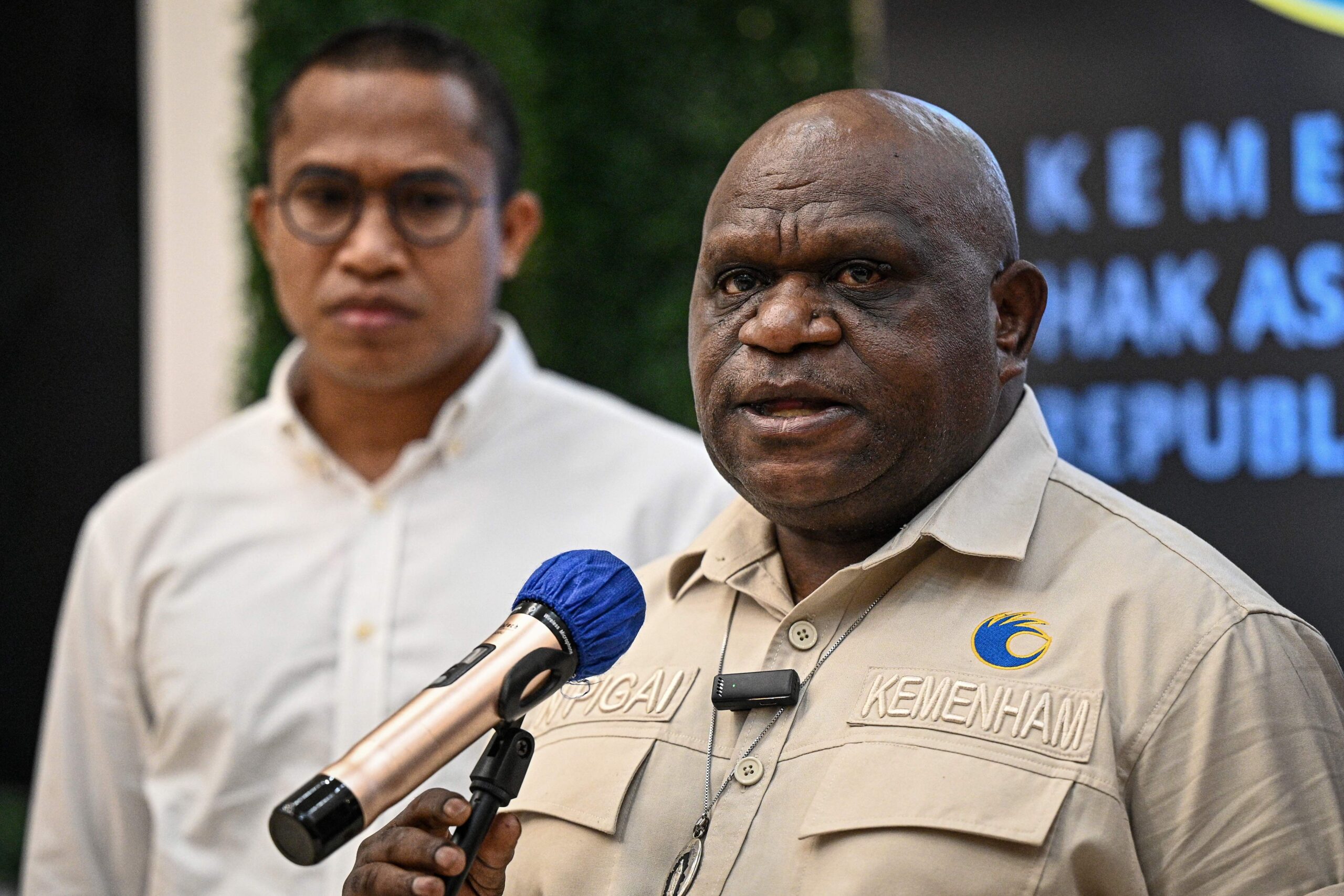 Pigai catrawarta Close up of a uniformed officer speaking into a handheld microphone at a press event with a second man in a light shirt visible in the blurred background
