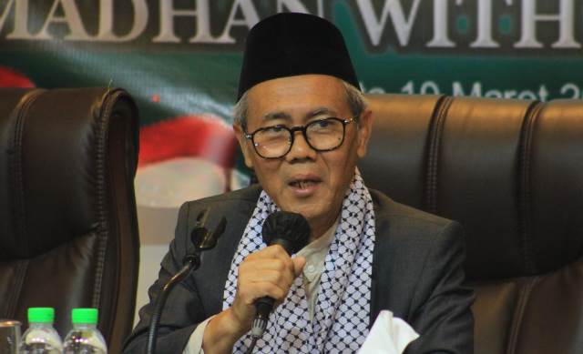 Man wearing a black cap and glasses, speaking into a handheld microphone at a panel.