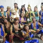 Group of performers in colorful traditional costumes posing for a group photo wearing ornate headpieces sashes and jewelry in a white backdrop setting