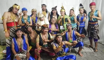Group of performers in colorful traditional costumes posing for a group photo, wearing ornate headpieces, sashes, and jewelry in a white backdrop setting.