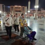 Beige uniformed men escort an elderly person in a wheelchair through a lit night time mosque courtyard
