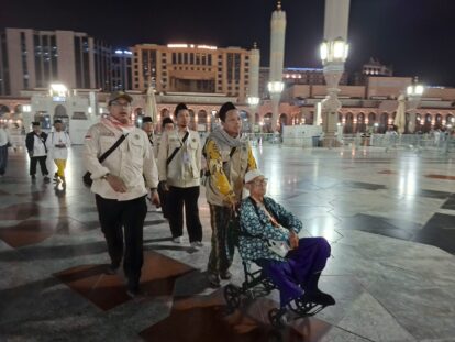Beige uniformed men escort an elderly person in a wheelchair through a lit night time mosque courtyard