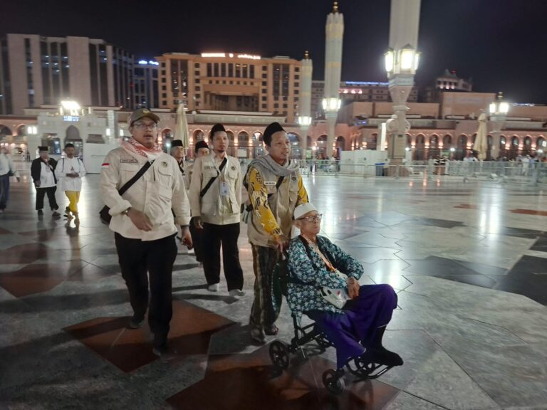 Beige-uniformed men escort an elderly person in a wheelchair through a lit night-time mosque courtyard.