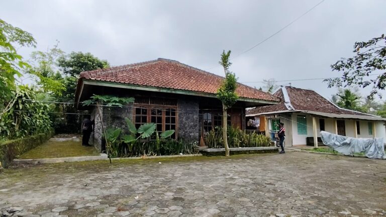 A village scene with two traditional tiled-roof houses, a cobblestone courtyard, and people near the entrance areas.