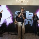 Lead singer wearing a bucket hat and sunglasses sings into a mic on a stage with bandmates playing guitars behind him chandeliers overhead